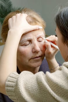 One young Caucasian girl cosmetologist draws a contour with a white marker on the left eyebrow of an elderly beautiful woman with her eyes closed, sitting in a home beauty salon, top side close-up view.