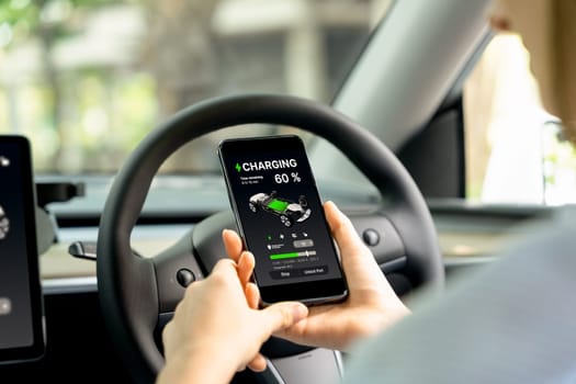 Young woman inside the vehicle, checking EV vehicle's application on battery recharging electricity status display on smartphone screen in modern EV car on her road trip journey. Exalt