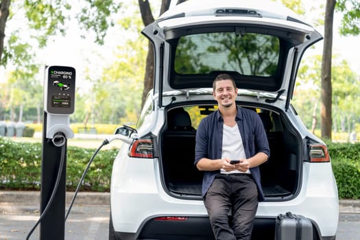 Man using smartphone online banking application to pay for electric car battery charging from EV charging station during vacation holiday road trip at national park or summer forest. Exalt