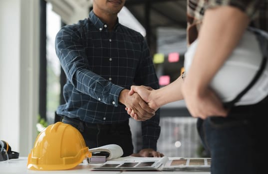 Two male engineers shaking hands in a modern office, collaborating on a construction project with safety helmet and design plans on the desk.