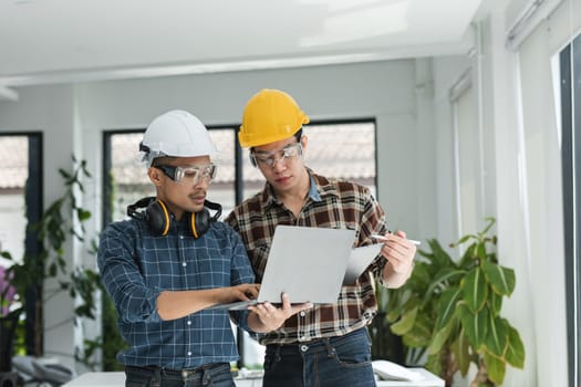 Two male engineers in a modern office, wearing safety helmets and protective glasses, collaborating on project plans using a laptop.