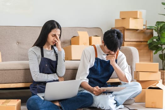 A couple feeling stressed while managing their small business's online sales, sitting on the floor with a laptop and tablet, surrounded by cardboard boxes.