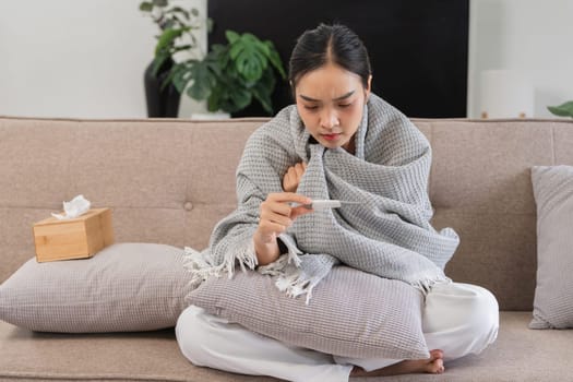 Woman with fever sitting on sofa, wrapped in blanket, checking thermometer, feeling unwell. Home interior, health and wellness concept.