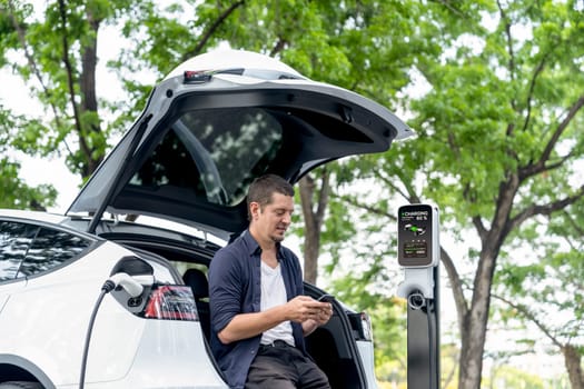 Man using smartphone online banking application to pay for electric car battery charging from EV charging station during vacation holiday road trip at national park or summer forest. Exalt