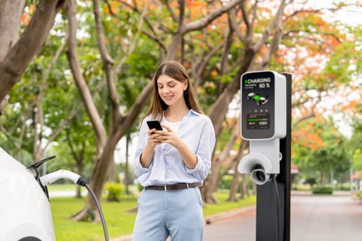 Young woman using smartphone online banking application to pay for electric car battery charging from EV charging station during autumn vacation holiday trip at national park or autumnal forest. Exalt