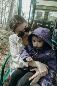 Portrait of one beautiful young Caucasian brunette girl in sunglasses and stylish clothes holds a baby girl in her arms, sitting in an open carriage of a tourist train, driving through a nature reserve on a spring sunny day in a public park, close-up side view.