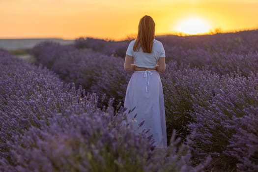 A woman stands in a field of lavender flowers. The sun is setting in the background, casting a warm glow over the scene