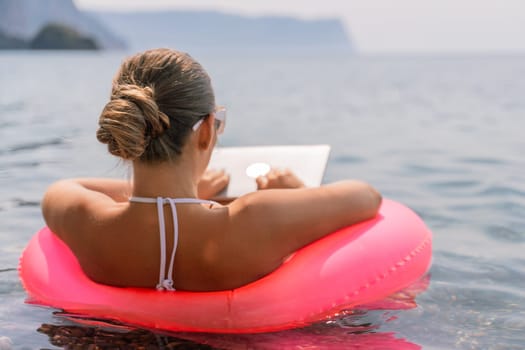 A woman is sitting on a pink inflatable raft in the ocean, reading a laptop. Concept of relaxation and leisure, as the woman enjoys her time by the water while working or browsing the internet