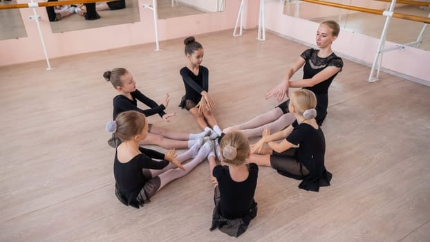 Caucasian woman and five little girls sit in a circle and do stretching at a ballet school