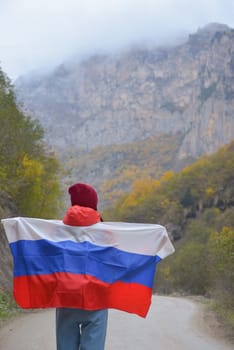 Woman traveler on a winding road in the mountains on a foggy autumn day with the flag of the Russian Federation on her shoulders.