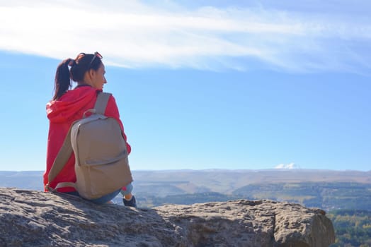 A girl tourist with a backpack sits on the top of a mountain and enjoys the beautiful views. A walking woman in an orange jacket rests on a rock, looking at the beautiful sunny landscape of the Caucasus Mountains