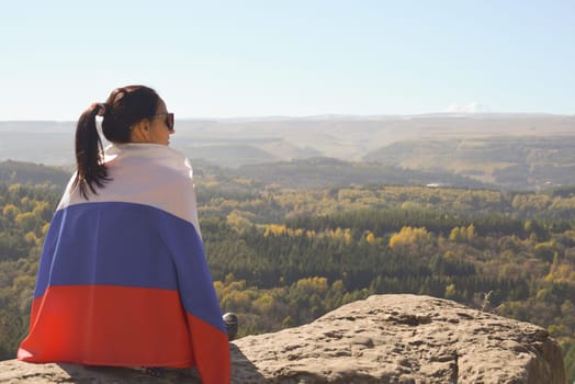 A girl tourist with a Russian flag on her shoulders sits on the top of a mountain and enjoys the beautiful views of the Caucasus Mountains