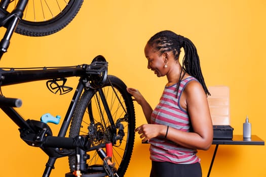 Active young woman does yearly maintenance and adjustment of bicycle tire in front of isolated yellow background. Athletic african american lady examining her bike wheel for recreational cycling.