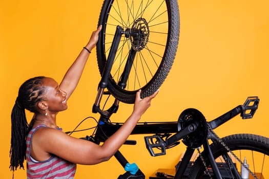 Sports-loving black woman carefully examines a bicycle, replacing a damaged rear tire. Healthy committed african american female cyclist safely reattaching bicycle wheel in front of yellow background.