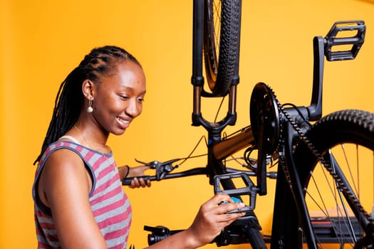 Dedicated black woman performing bike maintenance for leisure cycling. Healthy enthusiastic african american lady inspecting and fixing bike pedal doing annual adjustments against isolated background.