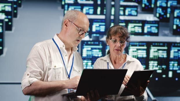 Focused IT inspectors looking around data center, using laptop and tablet to crosscheck disaster recovery plan and assess high tech establishment units in need of replacement, preventing perils