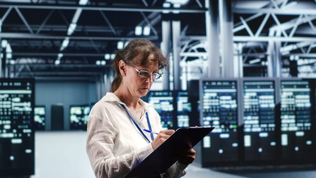 Aged IT admin in modern server room holding clipboard during data center maintenance, crossing off completed tasks. Trained specialist in high tech workspace finishing checkup on equipment