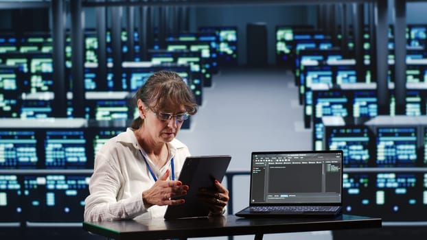 Senior computer scientist in server farm completing tasks on tablet while running scripts on laptop terminal. Expert amidst supercomputers manipulating code and improving cybersecurity