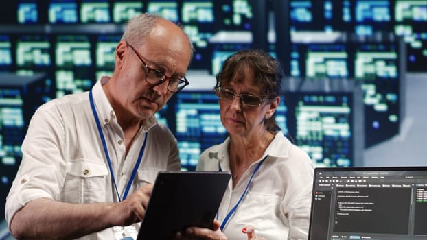 Tech savvy inspectors solving tasks on tablet while scripts are running on laptop terminal. Productive IT practitioners amidst servers, accessing databases and manipulating lines of code