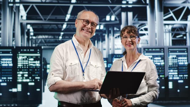 Coworkers in data center using laptop to monitor energy efficient server racks. Equipment using renewable sources employed to minimize consumption and reduce overall facility carbon footprint