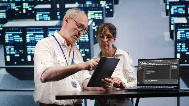 Hardworking engineers in high tech facility, solving tasks on tablet while running scripts on laptop terminal. Dilligent technicians amidst blade servers, accessing databases and manipulating code