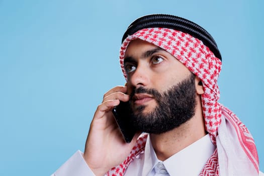 Muslim man wearing islamic headscarf looking up while talking on smartphone in studio. Arab person dressed in ghutra headdress having conversation on mobile phone closeup