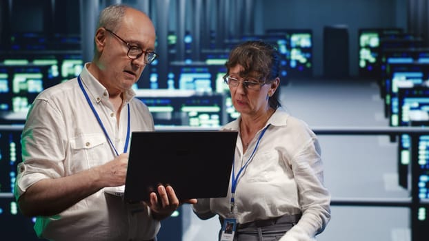 Adept mechanics looking around high tech data center, using laptop to crosscheck disaster recovery plan and assess server clusters in need of replacement, preventing pitfalls