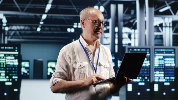 Focused IT professional navigating network of blade servers in industrial hub. Serviceman with laptop ensuring optimum operations, doing optimizations and maintenance in data center