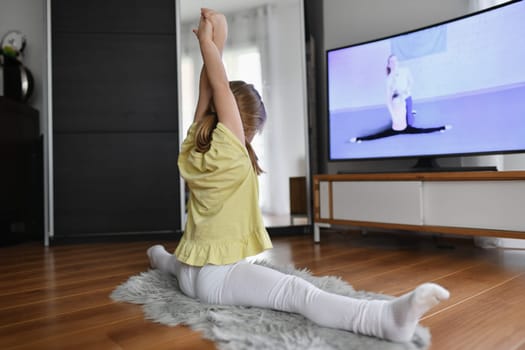 The girl sits on the splits and does gymnastics at home while watching a blogger on TV