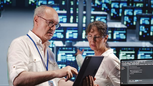 Organized admins in high tech workspace, completing tasks on tablet while running scripts on laptop terminal. Focused specialists amidst server clusters, accessing databases and manipulating code