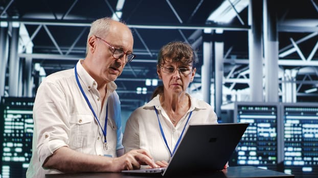 Octogenarian IT team looking around high tech data center, using laptop to crosscheck disaster recovery plan and assess server devices in need of replacement, preventing bugs