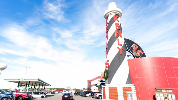 Tall black-and-white lighthouse sign with red accents in front of a historic building, under a clear blue sky with white clouds. Gas pumps and cars in parking lot by the lighthouse.