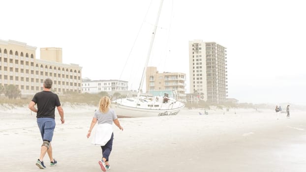 A man and woman are walking away from a beached sailboat on a foggy day. The image is taken from a low angle, making the boat look larger than life. There are buildings in the background.