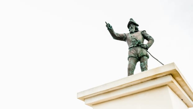 A bronze statue of Christopher Columbus stands atop a marble pedestal, pointing at the horizon with one hand while holding a map in the other. The statue is set against a solid white background.