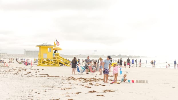 A diverse group of people, adults and kids, having fun at the beach near a lifeguard tower on an overcast day. Theyre enjoying the warm weather and creating happy memories together.