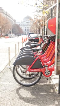 A vibrant row of red bicycles available for rent in the city, lined up near the sidewalk on a sunny day. Background shows blurred cars on the street and a pedestrian walking by.