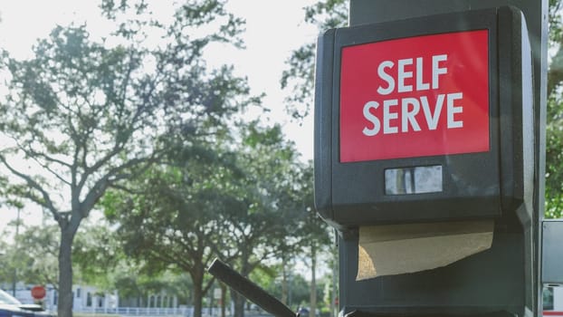 A tranquil self-serve gas station sign surrounded by lush green trees. The red sign with white lettering stands out, blending nature and modern convenience harmoniously.