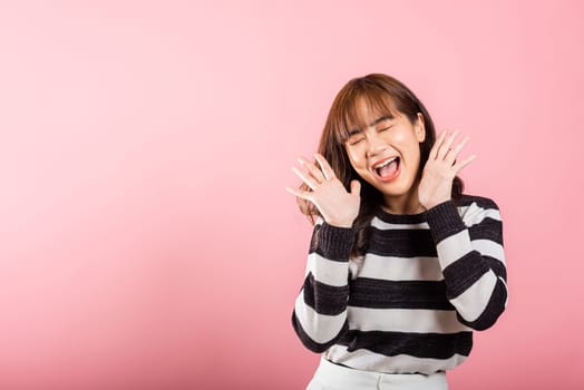 In a studio shot on a pink background, a jubilant Asian woman raises her fists in celebration, expressing success and happiness with a bright smile.