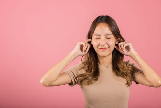 Woman bad mood pain her closed ears with finger and close eyes, Asian beautiful young female covers ears for loud noise, studio shot isolated on pink background with copy space, bad pressure