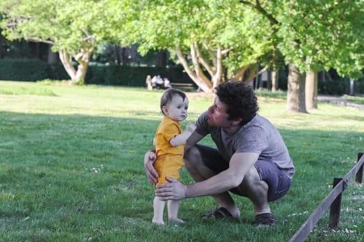 Caucasian young dad in a gray t-shirt, shorts with curly brown hair holds his little daughter in yellow overalls, standing barefoot on the grass, holding a daisy and reaching out to gently kiss her on the cheek in a public park, close-up side view. The concept of fatherhood, dads, family vacation, spring walk, first steps, happy childhood, PARKS and REC.