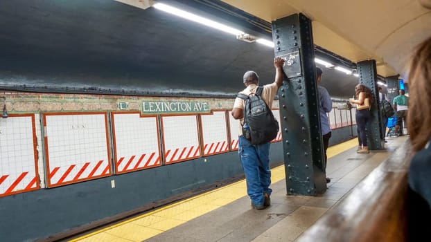 A young man waits on the platform at Lexington Avenue subway station in NYC. He wears jeans, a t-shirt, and carries a backpack. The station is old, dirty, with graffiti, trash.