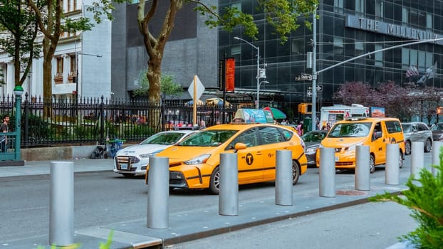 A yellow taxi cab drives through a busy city street with skyscrapers and office buildings in the background. Taxi cab is centered, background slightly blurred. Low angle shot, focused on taxi cab.