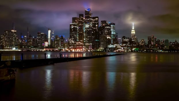 A captivating night view of New York City with sparkling lights reflected in the river. The dark sky and tall buildings create a stunning cityscape, showcasing the vibrant essence of the metropolis.