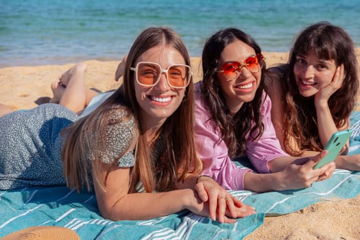 Group of smiling multiethnic women wearing hats and sunglasses lying on a towel on the sand enjoying vacation. Beautiful and cheerful Gen Z girls pose looking at the camera