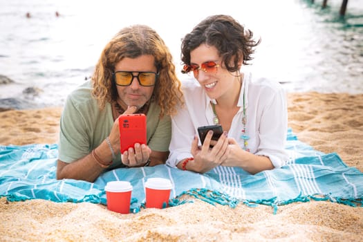 Middle-aged man and woman sitting on the beach with surprised face, browsing smartphone apps. Concept:Holidays y Technology