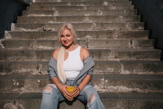 A blonde woman is sitting on a set of stairs, holding a cup of coffee. She is wearing a white tank top and jeans
