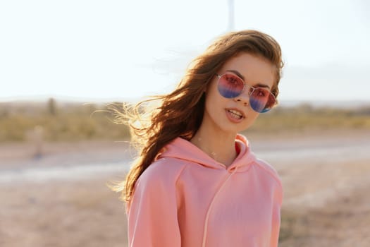 Stylish young woman in pink hoodie and sunglasses walking on desert road under sunny sky