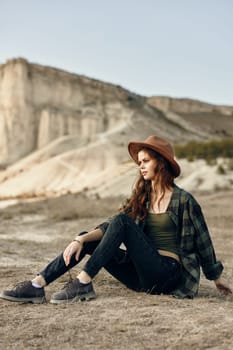 Serene young woman in floppy hat sitting peacefully in desert with majestic mountains in the distance