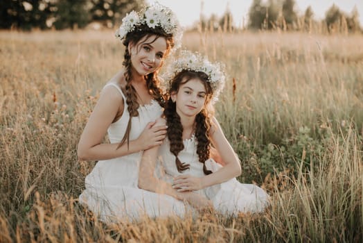 Happy mom and daughter smiling and hugging in field in the summer. Sisters in white dresses with floral wreaths and braids in boho style. Added a small grain simulation of film photography