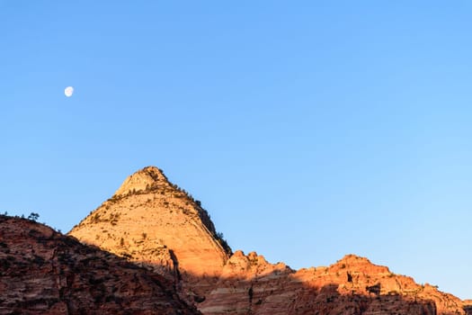 View of glowing canyon walls, showcasing bright sunlight on the rocky peaks and a visible moon against a clear blue sky. Serene landscape captures the beauty of nature - Zion National Park, Utah, USA